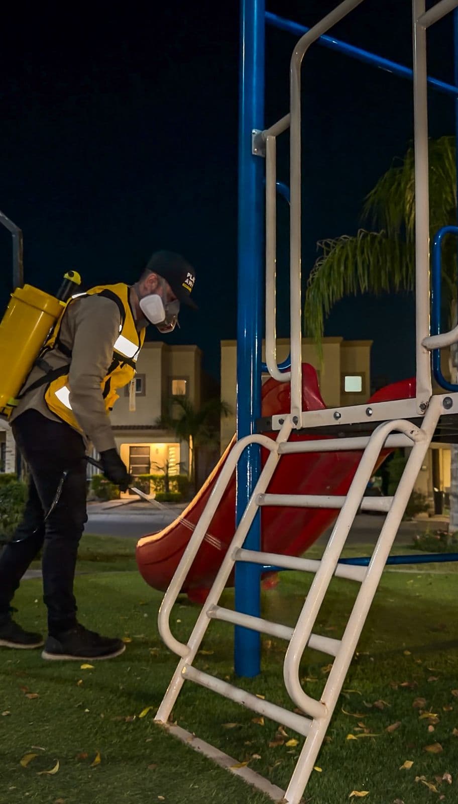Técnico de Plagaway fumigando de noche el área de juegos infantiles de un fraccionamiento, junto a una resbaladilla roja
